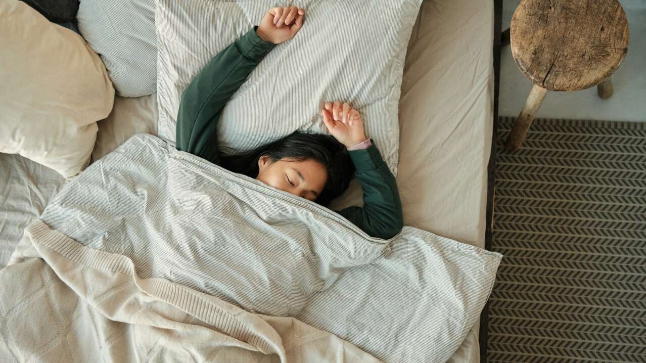 Young woman stretching in bed under a soft blanket, surrounded by pillows and a side table.